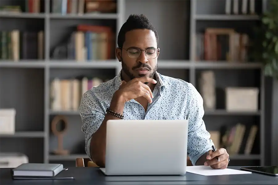 African American male author looking at laptop screen, touching chin, pondering ideas inside his home office