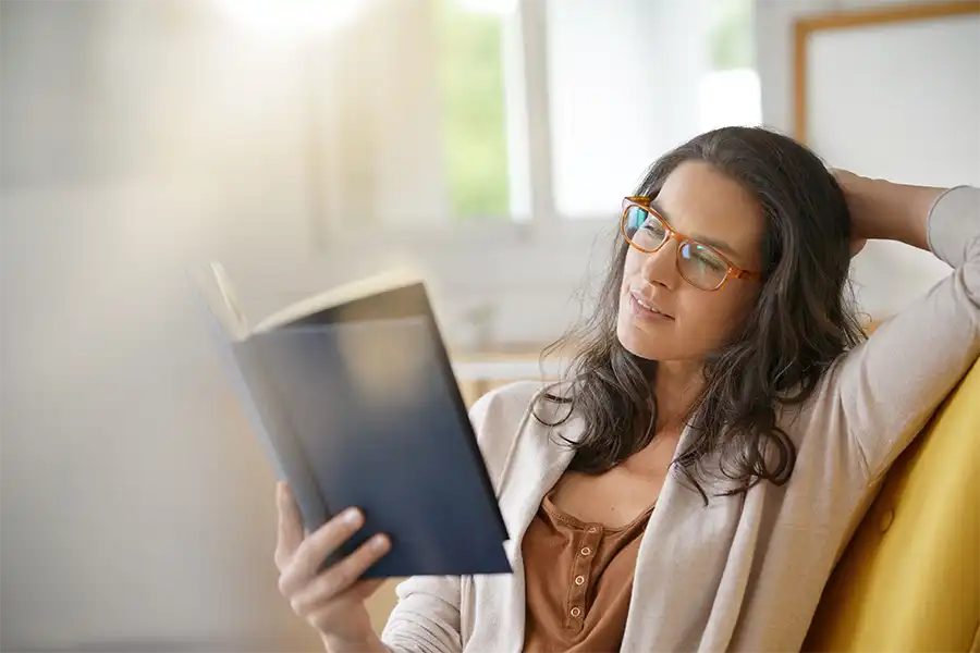A woman with long brown hair, glasses and a brown shirt with a cream sweater reading a book on her couch in her living room